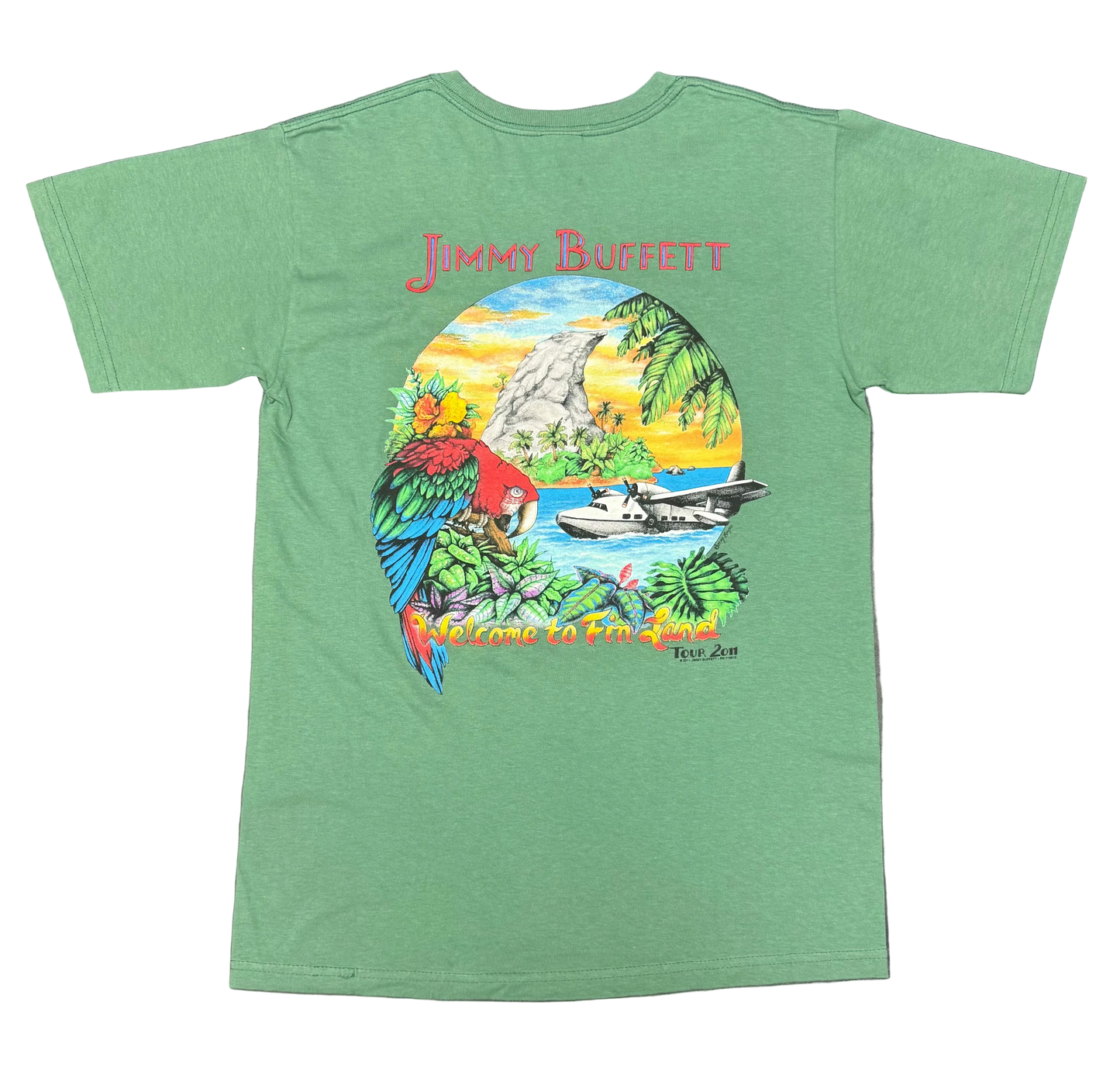 Back of a green short sleeve Jimmy Buffett tee featuring large red “Jimmy Buffett” lettering above a tropical island scene. The artwork includes a red, blue, and green macaw, lush foliage, a seaplane on the water, a fin shaped rock formation, and a sunset sky. “Welcome to Fin Land Tour 2011” appears along the bottom.