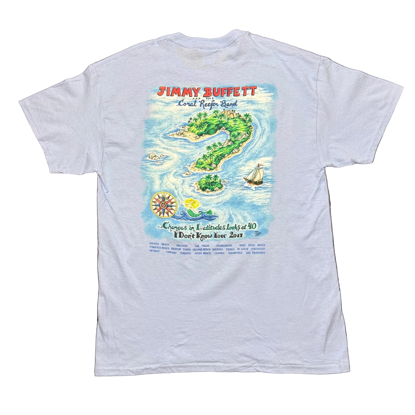 Back of a light blue short sleeve Jimmy Buffett tee featuring a large tropical island scene surrounded by blue water, with a sailboat on the right and a compass graphic near the lower left. “Jimmy Buffett and the Coral Reefer Band” appears across the top, with “Changes in Latitudes Looks at 40” and “I Don’t Know Tour 2017” printed below, followed by a list of tour cities at the bottom.