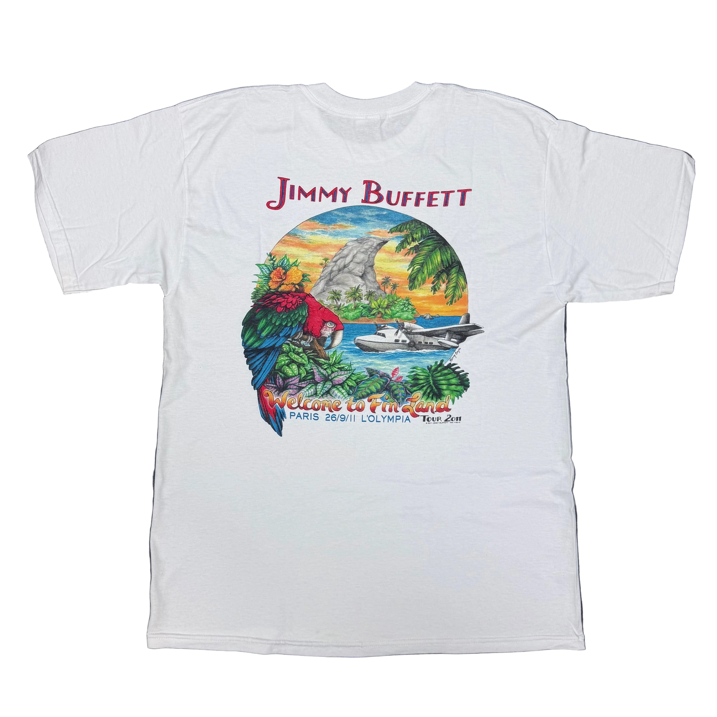 Back of a white short sleeve Jimmy Buffett tee featuring large red “Jimmy Buffett” lettering above a tropical island scene. The artwork includes a red, blue, and green macaw, lush foliage, a seaplane on the water, a fin shaped rock formation, and a sunset sky. “Welcome to Fin Land” appears along the bottom with “Paris 26/9/11 L’Olympia Tour 2011” beneath.