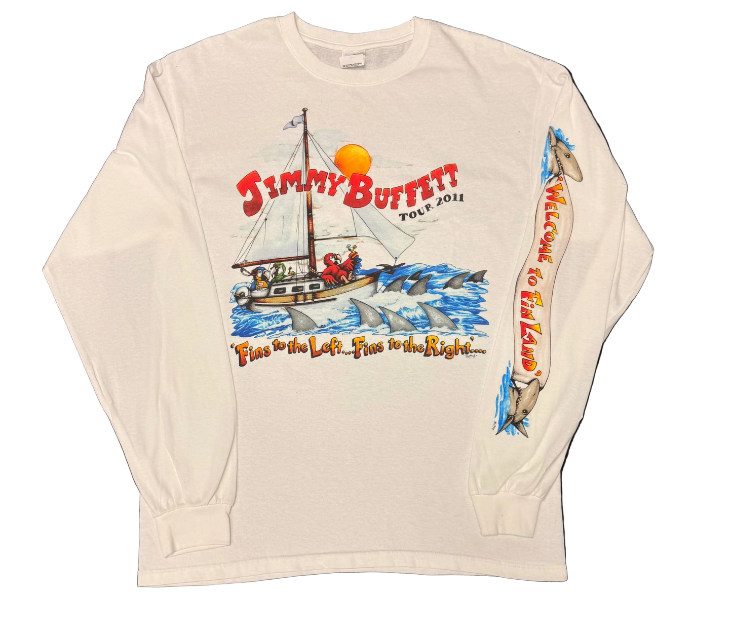 White long sleeve Jimmy Buffett tee featuring a sailboat surrounded by shark fins in blue ocean waves. Red “Jimmy Buffett Tour 2011” lettering appears above, with “Fins to the Left, Fins to the Right…” below. One sleeve features shark graphics and vertical “Welcome to Fin Land” lettering.