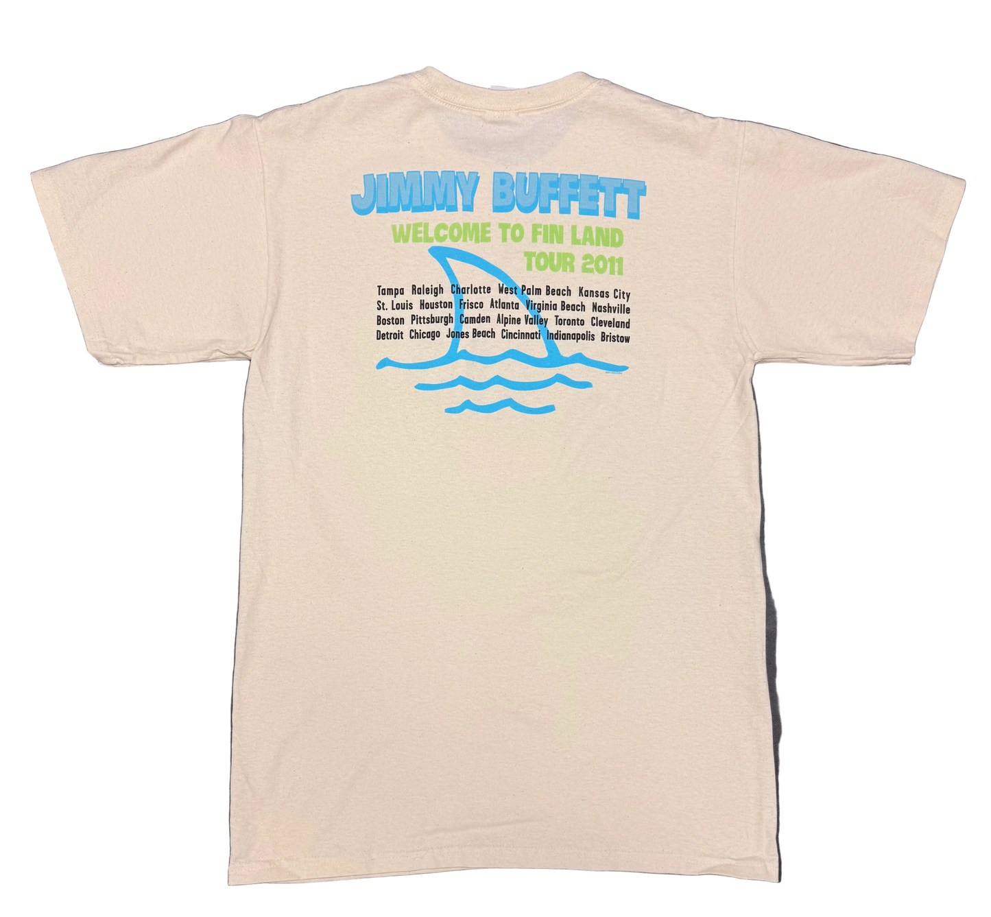 Back of a natural off white short sleeve Jimmy Buffett tee featuring large blue “Jimmy Buffett” lettering above green “Welcome to Fin Land Tour 2011” text. A large blue shark fin and wave graphic appears behind a list of tour cities printed in black.
