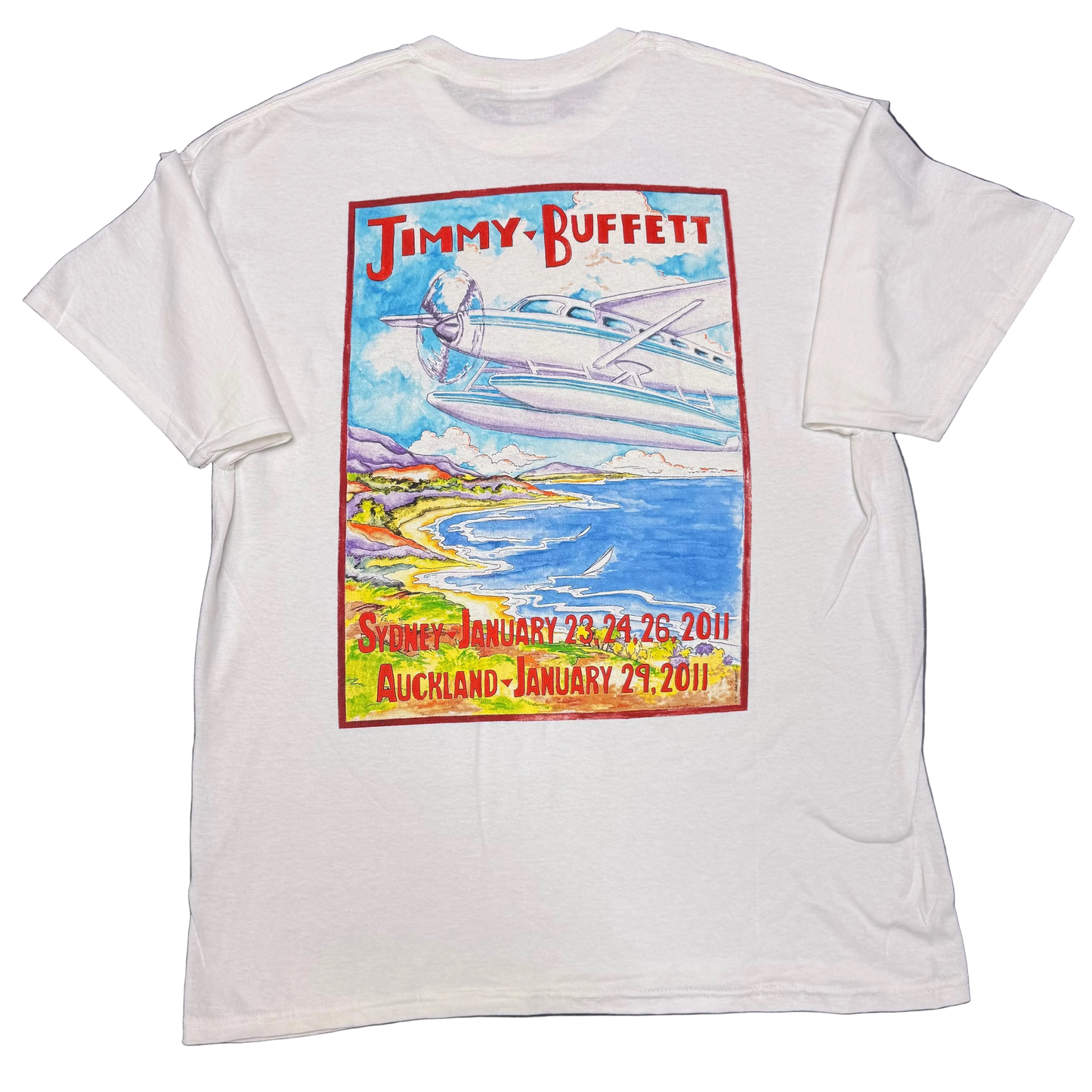 Back of a white short sleeve Jimmy Buffett tee featuring a large colorful coastal scene with a seaplane flying over blue water and shoreline. Red “Jimmy Buffett” lettering appears across the top, with Sydney dates January 23, 24, and 26, 2011 and Auckland January 29, 2011 printed along the bottom.