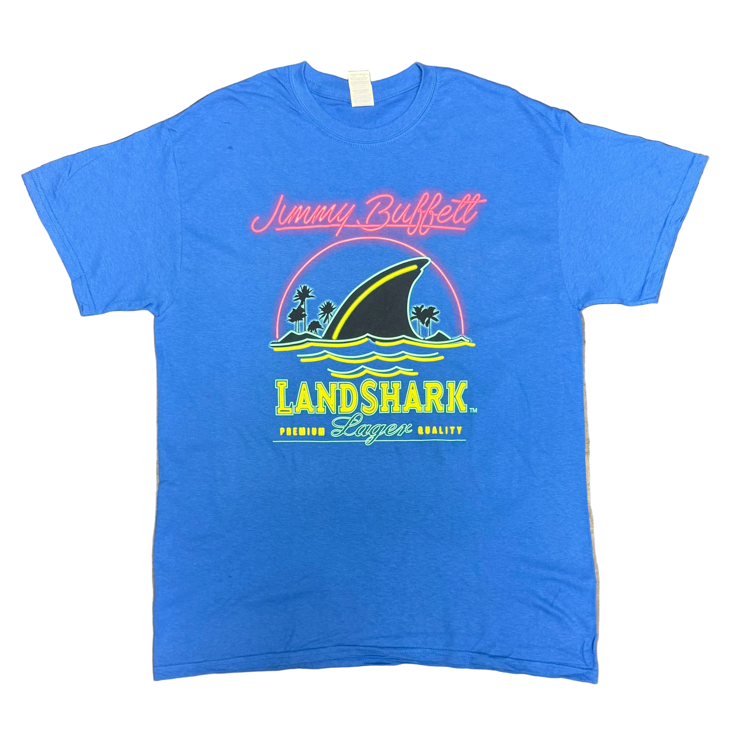 Royal blue short sleeve Jimmy Buffett tee featuring pink script “Jimmy Buffett” lettering above a large LandShark Lager graphic. The design includes a black shark fin cutting through blue waves, small palm trees, and bright yellow, green, and pink accents.