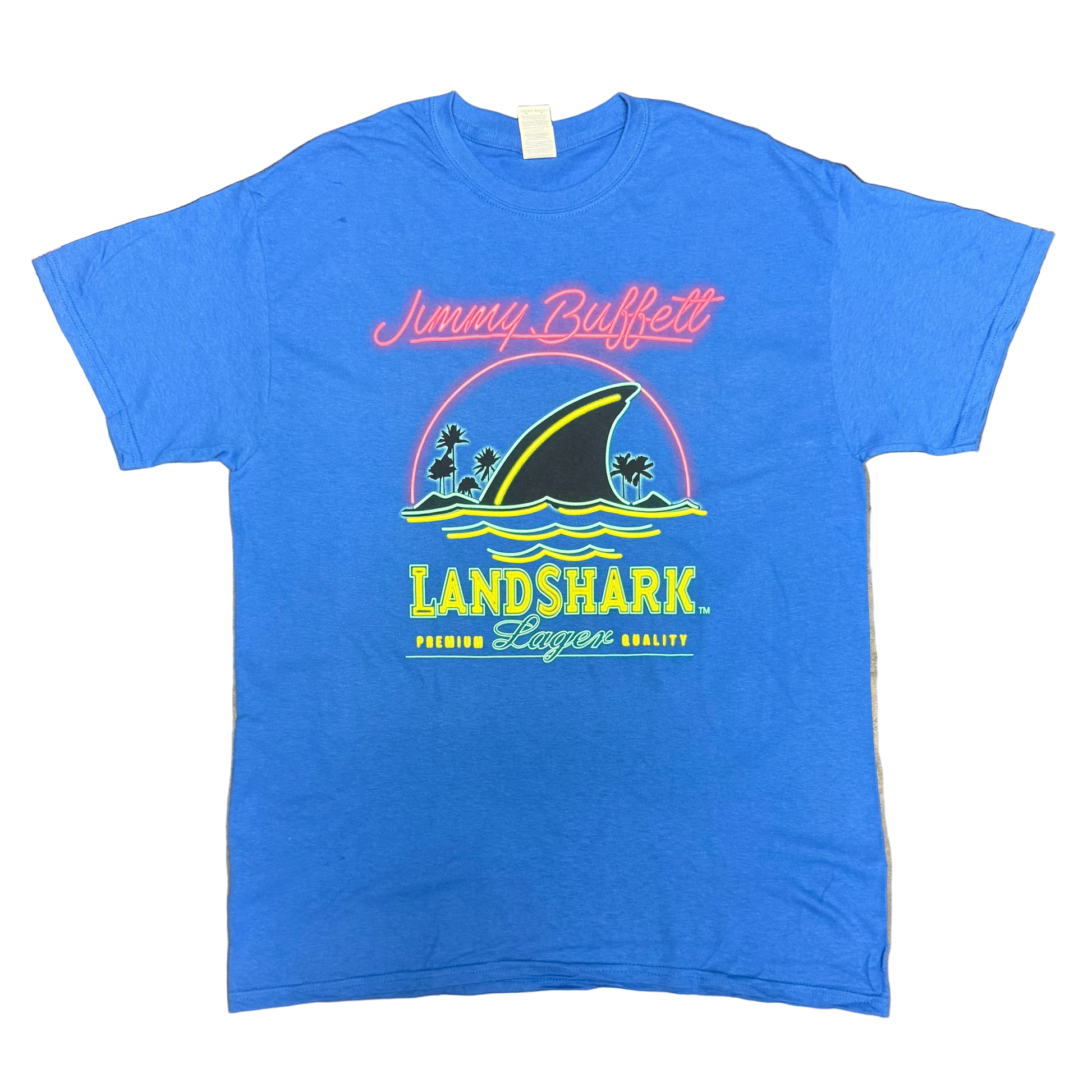Royal blue short sleeve Jimmy Buffett tee featuring pink script “Jimmy Buffett” lettering above a large LandShark Lager graphic. The design includes a black shark fin cutting through blue waves, small palm trees, and bright yellow, green, and pink accents.