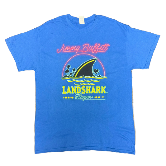 Royal blue short sleeve Jimmy Buffett tee featuring pink script “Jimmy Buffett” lettering above a large LandShark Lager graphic. The design includes a black shark fin cutting through blue waves, small palm trees, and bright yellow, green, and pink accents.
