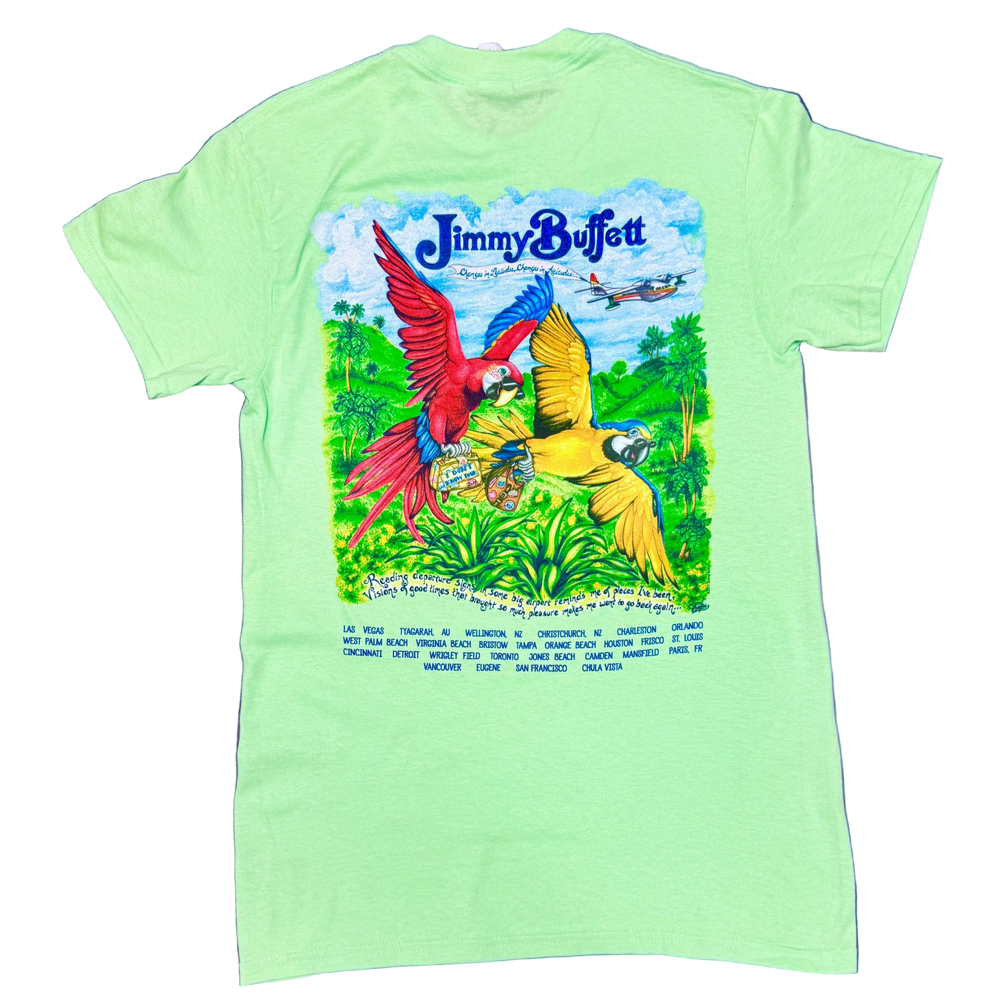 Back of a light green short sleeve Jimmy Buffett tee featuring large blue “Jimmy Buffett” lettering above a tropical scene with two colorful macaws flying over lush green foliage. A seaplane flies in the background, and a handwritten lyric line appears near the bottom above a list of 2017 tour cities printed in blue.